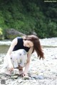 A woman crouches down on a rocky beach next to a river.